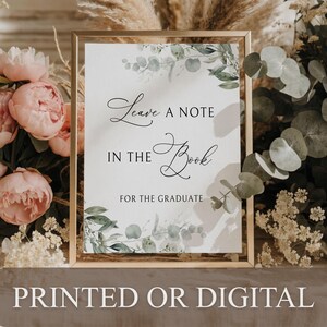 May include: A vertical rectangular sign in a thin gold frame featuring the black text Leave a note in the book for the graduate. The white background is decorated with green eucalyptus leaves and botanical illustrations at the top and bottom.