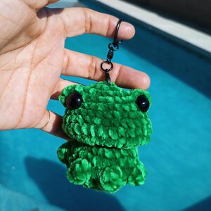 May include: A green crocheted frog keychain with black eyes and a black metal clasp. The frog is made of a soft, textured yarn and has a rounded body and head. The keychain is held up against a blue background.
