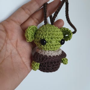 Puede incluir: Un personaje verde y marr&oacute;n tejido a ganchillo con grandes ojos negros y un cord&oacute;n marr&oacute;n. El personaje tiene orejas grandes y viste un atuendo marr&oacute;n y beige. El art&iacute;culo est&aacute; hecho a mano.