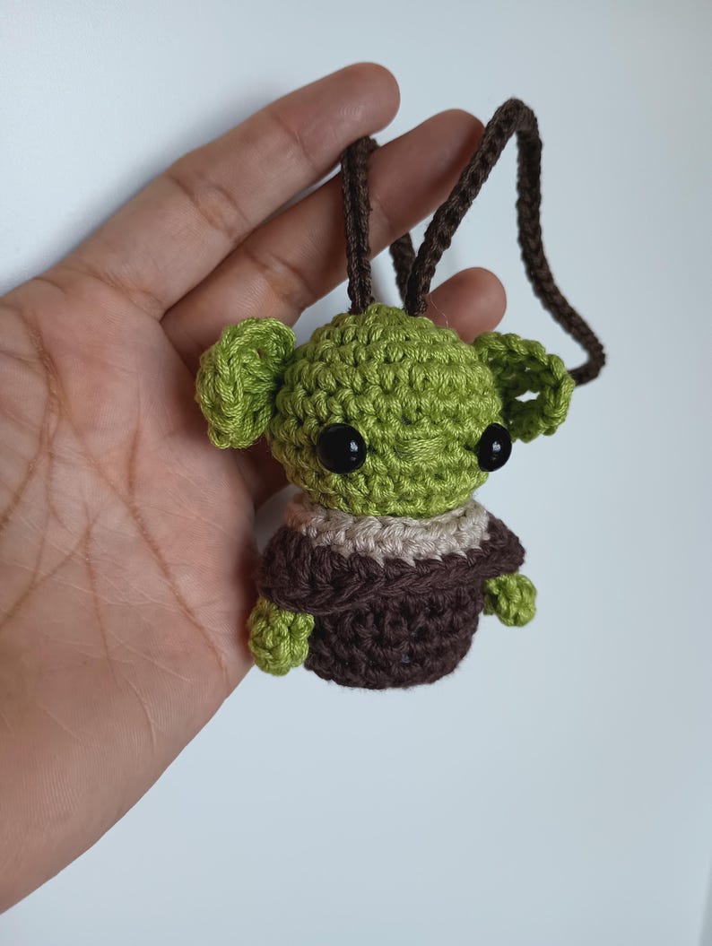 Puede incluir: Un personaje verde y marr&oacute;n tejido a ganchillo con grandes ojos negros y un cord&oacute;n marr&oacute;n. El personaje tiene orejas grandes y viste un atuendo marr&oacute;n y beige. El art&iacute;culo est&aacute; hecho a mano.
