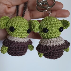 Puede incluir: Dos figuras de extraterrestres verdes de ganchillo con ojos negros y t&uacute;nicas marrones. Una est&aacute; unida a un cord&oacute;n marr&oacute;n, la otra a un llavero plateado. Las figuras tienen orejas grandes y est&aacute;n hechas a mano.