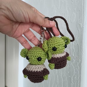 Puede incluir: Dos figuras de ganchillo verdes con aspecto de extraterrestre, con grandes ojos negros, t&uacute;nicas marrones y cuellos beige. Est&aacute;n unidas a llaveros y un cord&oacute;n marr&oacute;n. Las figuras se sostienen sobre un fondo blanco.