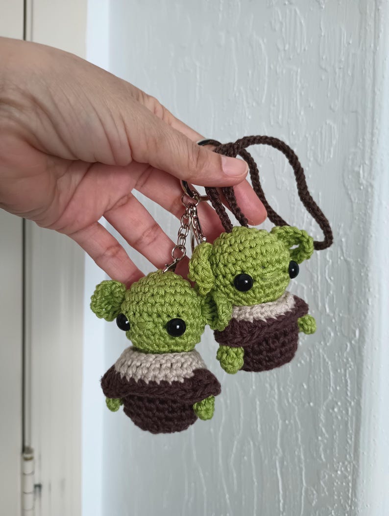 Puede incluir: Dos figuras de ganchillo verdes con aspecto de extraterrestre, con grandes ojos negros, t&uacute;nicas marrones y cuellos beige. Est&aacute;n unidas a llaveros y un cord&oacute;n marr&oacute;n. Las figuras se sostienen sobre un fondo blanco.