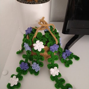 May include: A handmade crochet hanging basket with trailing vines and flowers. The basket is tan, with green vines and white and purple flowers. The hanging cord is tan. The basket is sitting on a white surface.