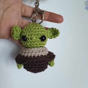Puede incluir: Un llavero verde tejido a mano que representa un personaje con orejas grandes y ojos negros. El personaje lleva un atuendo marr&oacute;n y beige. El llavero tiene un cierre y un anillo plateados.