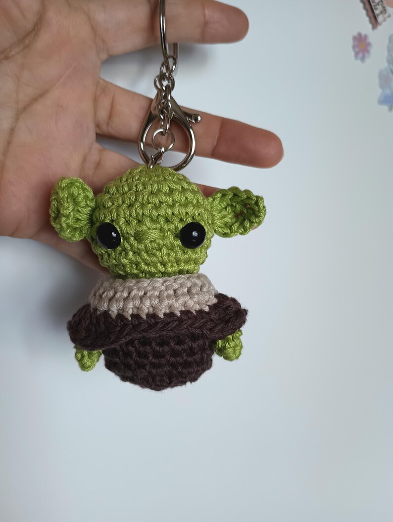 Puede incluir: Un llavero verde tejido a mano que representa un personaje con orejas grandes y ojos negros. El personaje lleva un atuendo marr&oacute;n y beige. El llavero tiene un cierre y un anillo plateados.