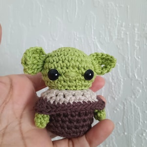 Puede incluir: Peque&ntilde;a figura de ganchillo de un personaje con orejas grandes, ojos negros y tez verde. La figura lleva un atuendo marr&oacute;n y beige. El art&iacute;culo hecho a mano se sostiene en una mano.