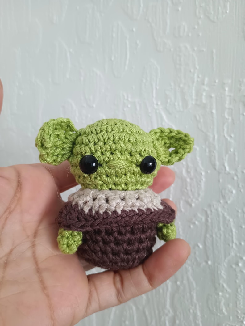 Puede incluir: Peque&ntilde;a figura de ganchillo de un personaje con orejas grandes, ojos negros y tez verde. La figura lleva un atuendo marr&oacute;n y beige. El art&iacute;culo hecho a mano se sostiene en una mano.