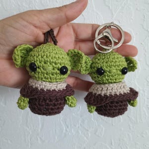 Puede incluir: Dos llaveros de ganchillo con un personaje alien&iacute;gena verde con orejas grandes y ojos negros. El personaje lleva un atuendo marr&oacute;n y beige. Un llavero tiene un lazo marr&oacute;n, el otro un cierre plateado.