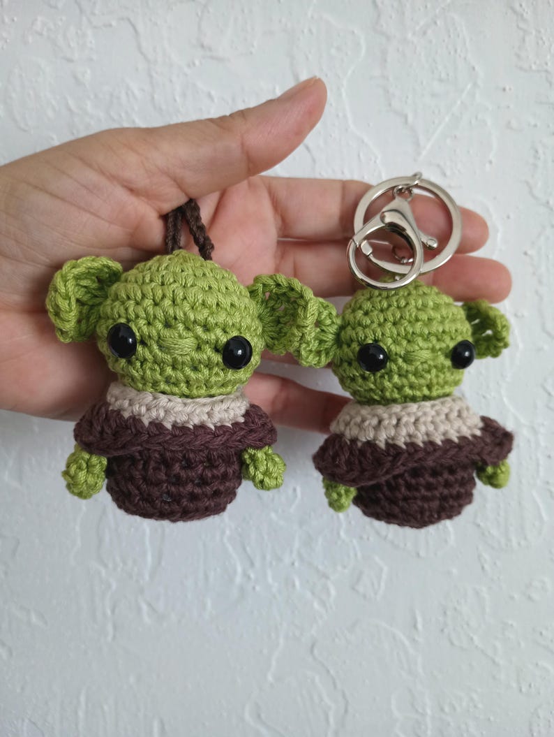 Puede incluir: Dos llaveros de ganchillo con un personaje alien&iacute;gena verde con orejas grandes y ojos negros. El personaje lleva un atuendo marr&oacute;n y beige. Un llavero tiene un lazo marr&oacute;n, el otro un cierre plateado.
