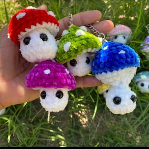 May include: A collection of handmade crochet mushroom keychains in various colors, including red, green, blue, and purple. Each mushroom has a white stem and black eyes, with white polka dots on the caps. The keychains are held in a hand against a backdrop of green grass.