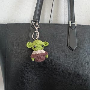 Puede incluir: Un llavero de ganchillo verde y marr&oacute;n hecho a mano con la forma de un personaje de ficci&oacute;n popular. El llavero est&aacute; unido a un llavero plateado y cuelga del asa de un bolso tote de cuero negro.