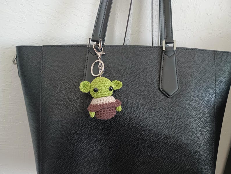 Puede incluir: Un llavero de ganchillo verde y marr&oacute;n hecho a mano con la forma de un personaje de ficci&oacute;n popular. El llavero est&aacute; unido a un llavero plateado y cuelga del asa de un bolso tote de cuero negro.