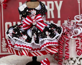 May include: A festive dog dress featuring a black fabric with a snowman print, layered with red and white striped and polka dot ruffles. A large red and white striped bow adorns the front. A matching bow is placed at the base.