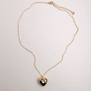 May include: A gold-toned necklace featuring a smooth, polished heart-shaped pendant suspended from a delicate cable chain. The chain includes an adjustable extender section with a small, singular charm at the end, providing a secure clasp closure.
