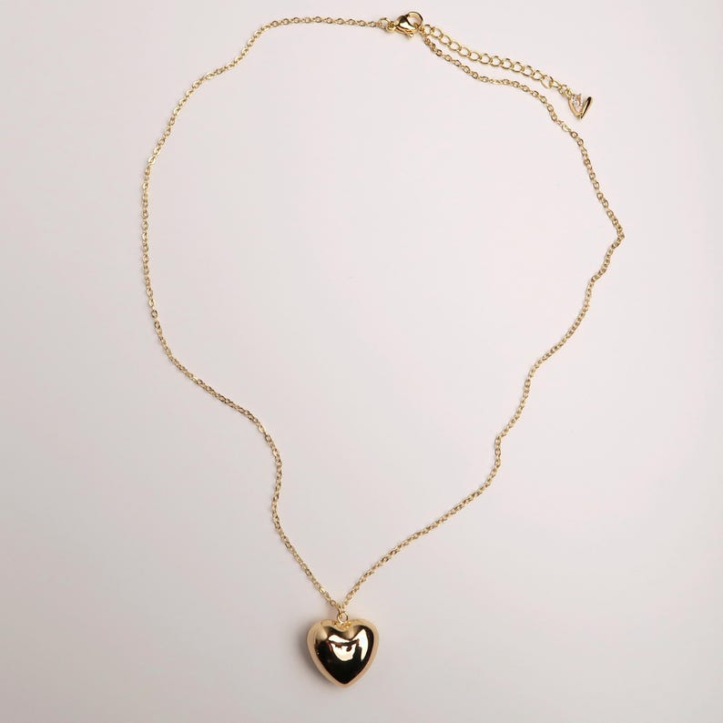 May include: A gold-toned necklace featuring a smooth, polished heart-shaped pendant suspended from a delicate cable chain. The chain includes an adjustable extender section with a small, singular charm at the end, providing a secure clasp closure.