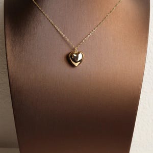 May include: A gold-toned necklace featuring a polished, smooth, three-dimensional heart-shaped pendant suspended from a delicate, thin metal chain. The heart pendant has a reflective, glossy surface that catches the light, creating a simple and clean aesthetic.