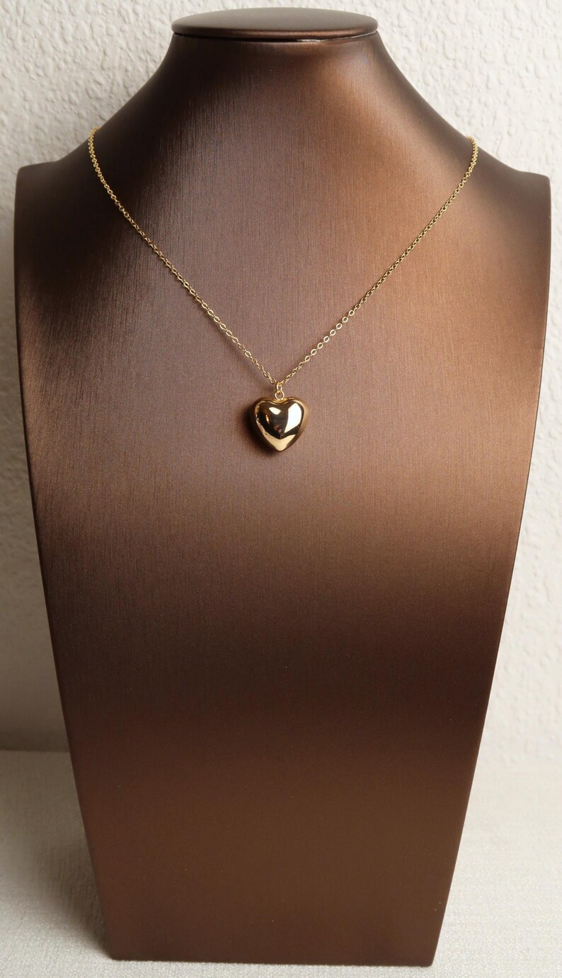 May include: A gold-toned necklace featuring a polished, smooth, three-dimensional heart-shaped pendant suspended from a delicate, thin metal chain. The heart pendant has a reflective, glossy surface that catches the light, creating a simple and clean aesthetic.
