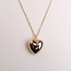 May include: A gold-toned necklace featuring a polished, smooth, puffed heart-shaped pendant suspended from a delicate matching gold-toned chain. The heart pendant has a reflective surface that catches light, creating bright highlights on its rounded form.