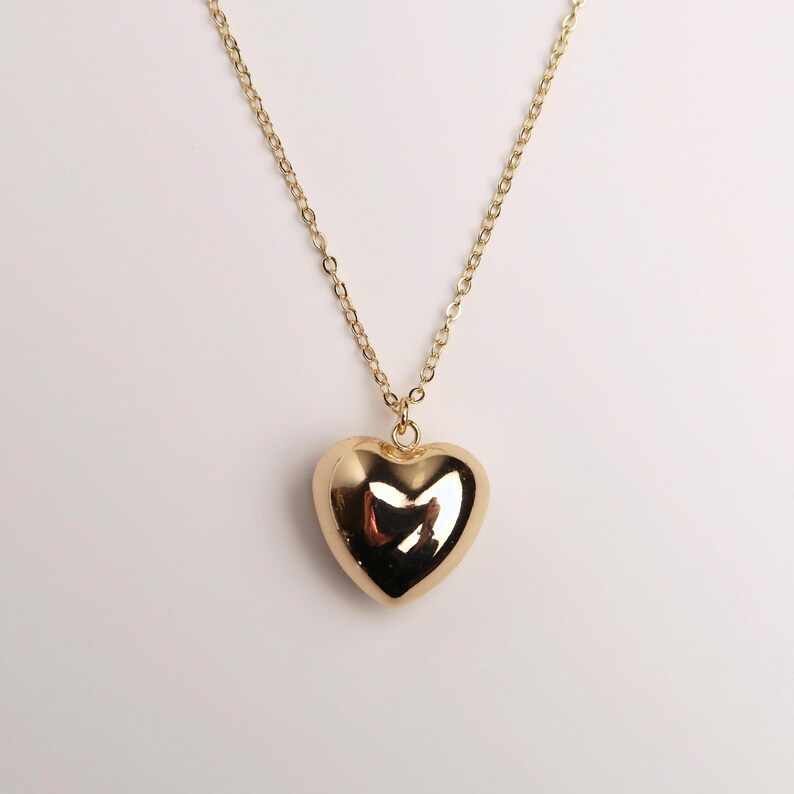 May include: A gold-toned necklace featuring a polished, smooth, puffed heart-shaped pendant suspended from a delicate matching gold-toned chain. The heart pendant has a reflective surface that catches light, creating bright highlights on its rounded form.
