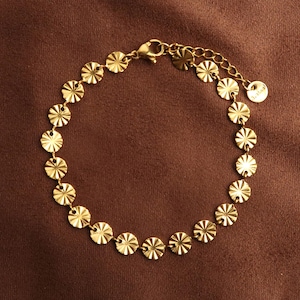 Puede incluir: Una pulsera dorada con una serie de pequeños abalorios circulares texturizados. Los abalorios están conectados por una delicada cadena y un cierre. La pulsera se muestra sobre un fondo marrón.