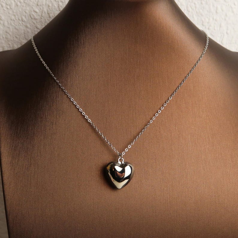 May include: A silver-toned necklace featuring a polished, smooth heart-shaped pendant suspended from a delicate, thin metal chain. The pendant has a reflective, metallic surface that catches the light, creating a simple and clean aesthetic.