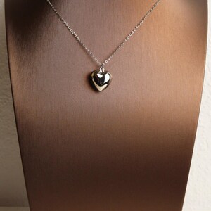 May include: A silver-toned necklace featuring a small, smooth, polished heart-shaped pendant suspended from a delicate, thin metal chain. The pendant has a reflective, metallic surface that catches the light, creating a simple and clean appearance.