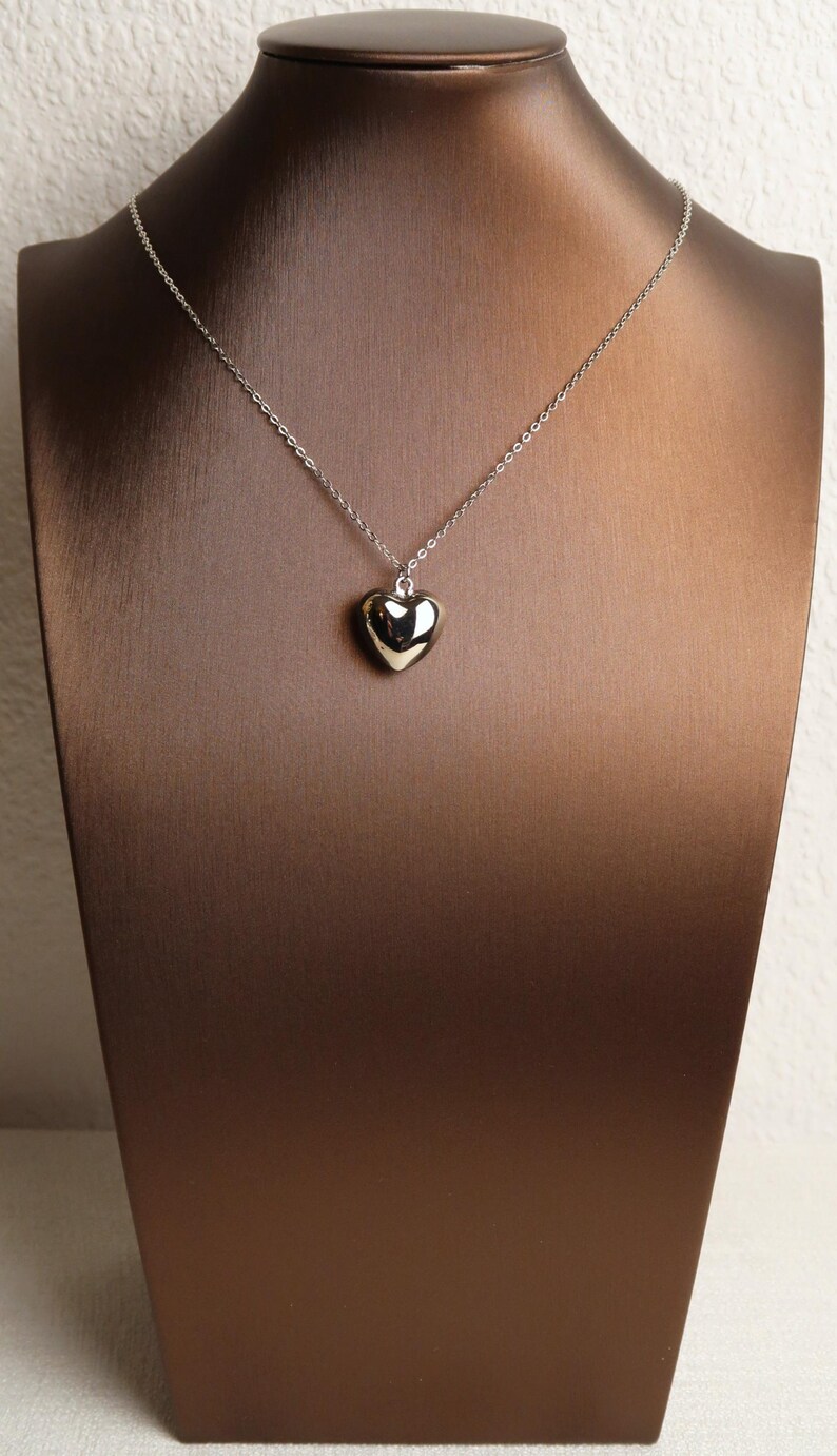 May include: A silver-toned necklace featuring a small, smooth, polished heart-shaped pendant suspended from a delicate, thin metal chain. The pendant has a reflective, metallic surface that catches the light, creating a simple and clean appearance.