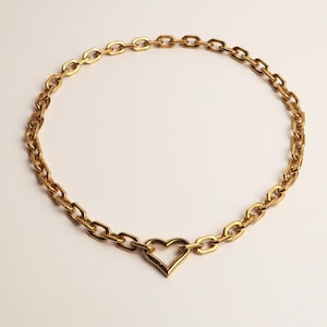 Puede incluir: Un collar de cadena en tono dorado que presenta una serie de eslabones ovalados alargados y uniformes conectados en un bucle continuo. El collar está centrado con un único eslabón en forma de corazón de marco abierto.