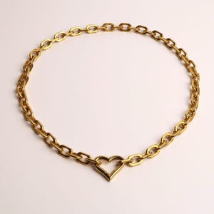 Könnte beinhalten: Eine goldfarbene Halskette mit einem herzförmigen Anhänger. Die Halskette besteht aus einer Reihe von ovalen Gliedern, die mit einem herzförmigen Anhänger verbunden sind. Der Schmuck wird auf weißem Hintergrund präsentiert.