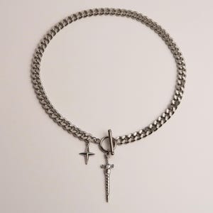 Puede incluir: Collar de cadena plateada con un diseño de eslabones de bordillo. El collar presenta un dije de cruz y un colgante de espada. El cierre es de estilo barra en T. La joya se exhibe sobre un fondo blanco.