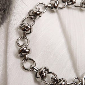 Puede incluir: Un brazalete de cadena de plata con un dise&ntilde;o anudado. El brazalete tiene un cierre de broche.