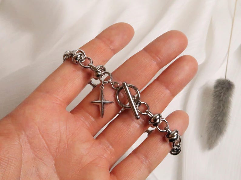 Puede incluir: Un brazalete plateado con cierre de palanca y un dije de estrella. El brazalete est&aacute; hecho de metal y tiene un dise&ntilde;o de cadena gruesa.
