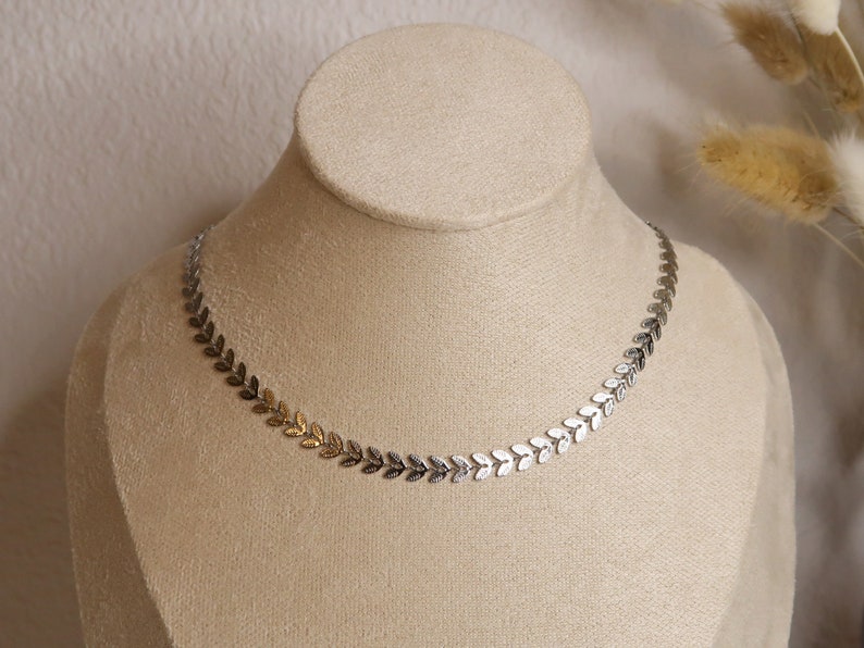 Puede incluir: Un collar de cadena de hojas plateadas y doradas en un expositor. El collar est&aacute; hecho de peque&ntilde;as hojas delicadas que est&aacute;n unidas entre s&iacute;.