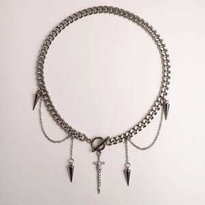Puede incluir: Collar de cadena plateada con cierre de palanca y colgantes colgantes. El collar presenta un diseño de cadena de eslabones gruesos, con tres colgantes en forma de cono y un colgante de espada. El collar se muestra sobre un fondo blanco.