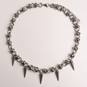 Puede incluir: Collar plateado con una cadena de anillos y cuentas interconectados. El collar tiene múltiples púas en forma de cono colgando de la parte inferior. El collar se muestra sobre un fondo blanco.