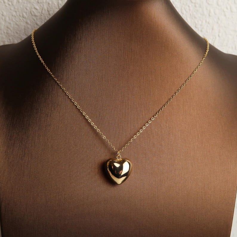May include: A gold-toned necklace featuring a delicate chain and a smooth, polished heart-shaped pendant. The pendant has a reflective surface that catches the light, suspended from a simple link chain that rests against the dark display form.
