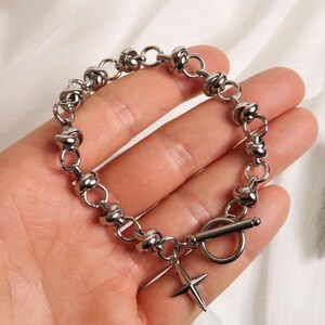 Puede incluir: Una pulsera de plata con un dise&ntilde;o de nudos y un peque&ntilde;o dije en forma de cruz. La pulsera tiene un cierre de palanca.