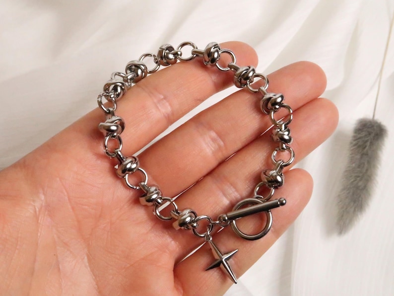 Puede incluir: Una pulsera de plata con un dise&ntilde;o de nudos y un peque&ntilde;o dije en forma de cruz. La pulsera tiene un cierre de palanca.