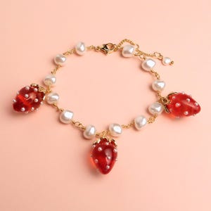 Peut inclure: Un bracelet doré orné de trois breloques fraises rouges avec des détails de graines blanches. Le bracelet est orné de perles blanches et possède une fermeture à fermoir mousqueton. Le fond est rose clair.