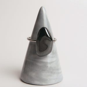 Könnte beinhalten: Ein Ring mit einem glatten, polierten, tropfenförmigen schwarzen Stein auf einem dünnen Metallband. Der Ring wird auf einem kegelförmigen Ständer aus grau-weißem Marmor präsentiert, was den Kontrast zwischen dem dunklen Stein und der Basis betont.