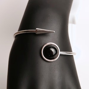 Może przedstawiać: Bransoletka na rękę w kolorze srebrnym z czarnym, okrągłym kamieniem i spiczastym detalem w kształcie stożka. Otwarta konstrukcja ułatwia noszenie. Nowoczesna biżuteria.
