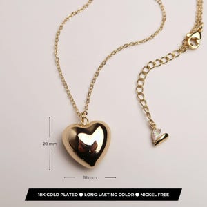 May include: A gold-plated necklace featuring a smooth, puffed heart pendant measuring 20 mm by 18 mm. The chain includes an adjustable extender with a small teardrop charm at the end. The text on the image states 18K GOLD PLATED, LONG-LASTING COLOR, NICKEL FREE.