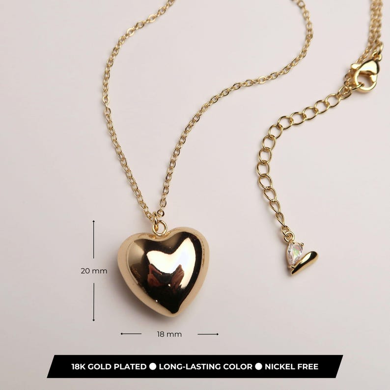 May include: A gold-plated necklace featuring a smooth, puffed heart pendant measuring 20 mm by 18 mm. The chain includes an adjustable extender with a small teardrop charm at the end. The text on the image states 18K GOLD PLATED, LONG-LASTING COLOR, NICKEL FREE.
