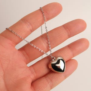 May include: A silver-toned metal necklace featuring a smooth, polished, three-dimensional heart-shaped pendant suspended from a delicate, thin link chain. The heart pendant has a reflective, mirror-like surface that catches light.