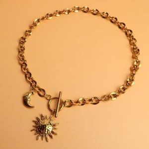 Puede incluir: Collar dorado con un diseño de cadena gruesa. El collar tiene un cierre de palanca e incluye dos colgantes: una luna creciente y un sol. La joya se muestra sobre un fondo color melocotón.