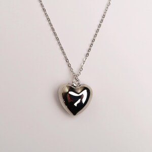 May include: A silver-toned necklace featuring a polished, puffed heart-shaped pendant suspended from a delicate, thin metal chain. The heart pendant has a smooth, reflective surface that catches the light, creating a bright, metallic appearance.