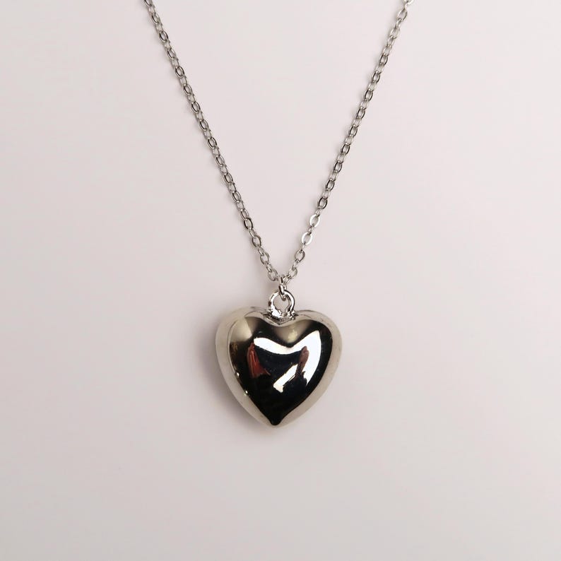 May include: A silver-toned necklace featuring a polished, puffed heart-shaped pendant suspended from a delicate, thin metal chain. The heart pendant has a smooth, reflective surface that catches the light, creating a bright, metallic appearance.