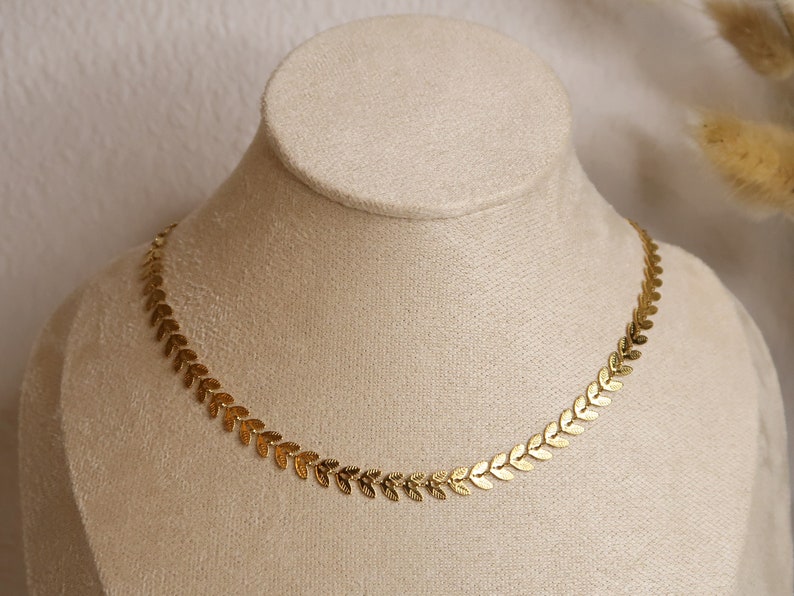 Puede incluir: Un collar de oro con una cadena delicada y peque&ntilde;os colgantes en forma de hoja. El collar se muestra en un maniqu&iacute; blanco.