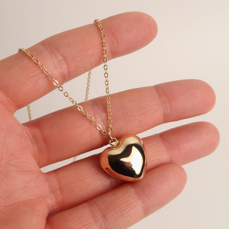 May include: A gold-toned necklace featuring a smooth, polished heart-shaped pendant suspended from a delicate, thin cable chain. The pendant has a reflective surface that catches the light, creating bright highlights across its rounded, puffed form.
